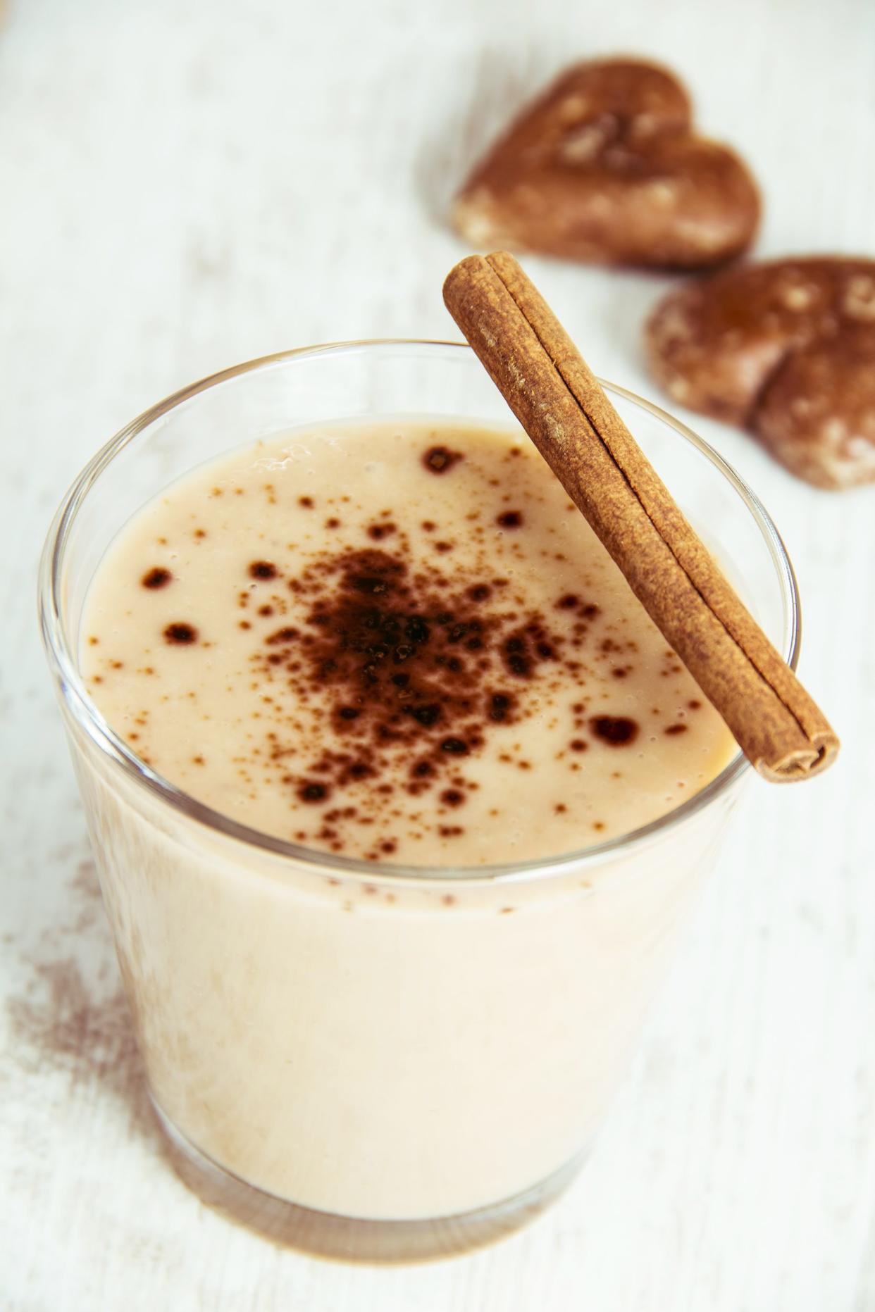 Top view of cinnamon bun smoothie in a small glass, with a cinnamon stick perched on top, on a white table with two heart cookies blurred in the background