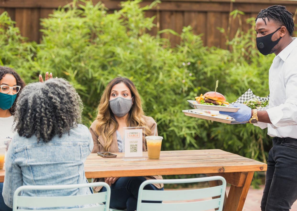 A male waiter brings customer's order to their table. He and the customers are wearing protective face masks amid the COVID-19 pandemic.