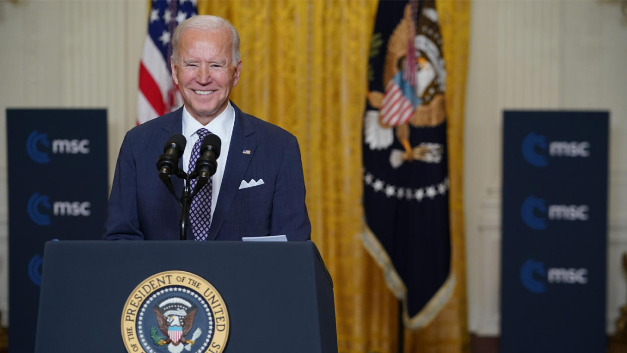 President Biden smiles behind a podium that bears two microphones and the presidential seal.
