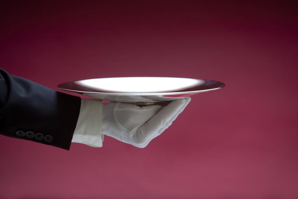 A gloved hand in a formal suit holds up an empty silver serving tray against a plain background