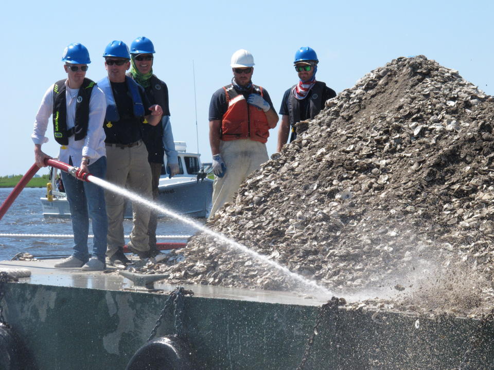 In this June 29, 2021 photo, New Jersey Environental Protection Commissioner Shawn LaTourette, left, uses a high-pressure hose to blast clam and oyster shells from a barge into the Mullica River in Port Republic, N.J. The shells are collected from restaurants in Atlantic City, dried, and placed into the river where they become the foundation for new oyster colonies as free-floating baby oysters attach to them and start to grow. Communities around the world are running similar shell recycling programs. (AP Photo/Wayne Parry)