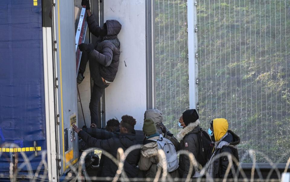 A group of migrants, some wearing facemasks look on as one of their number climbs into the back of a lorry, in front of a high fence