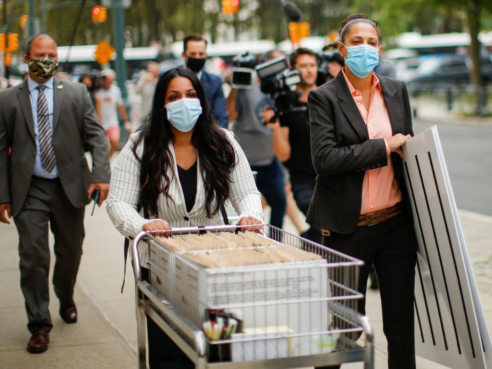 Prosecutors wearing facemasks push a cart with white cardboard boxes of evidence along a sidewalk