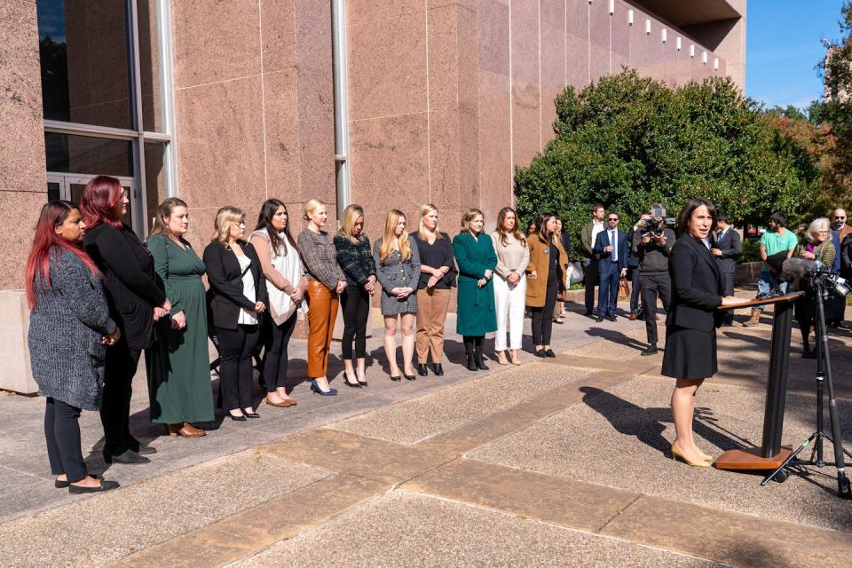 A woman in a black outfit stands at a podium in front of a long row of women who stand looking forward.