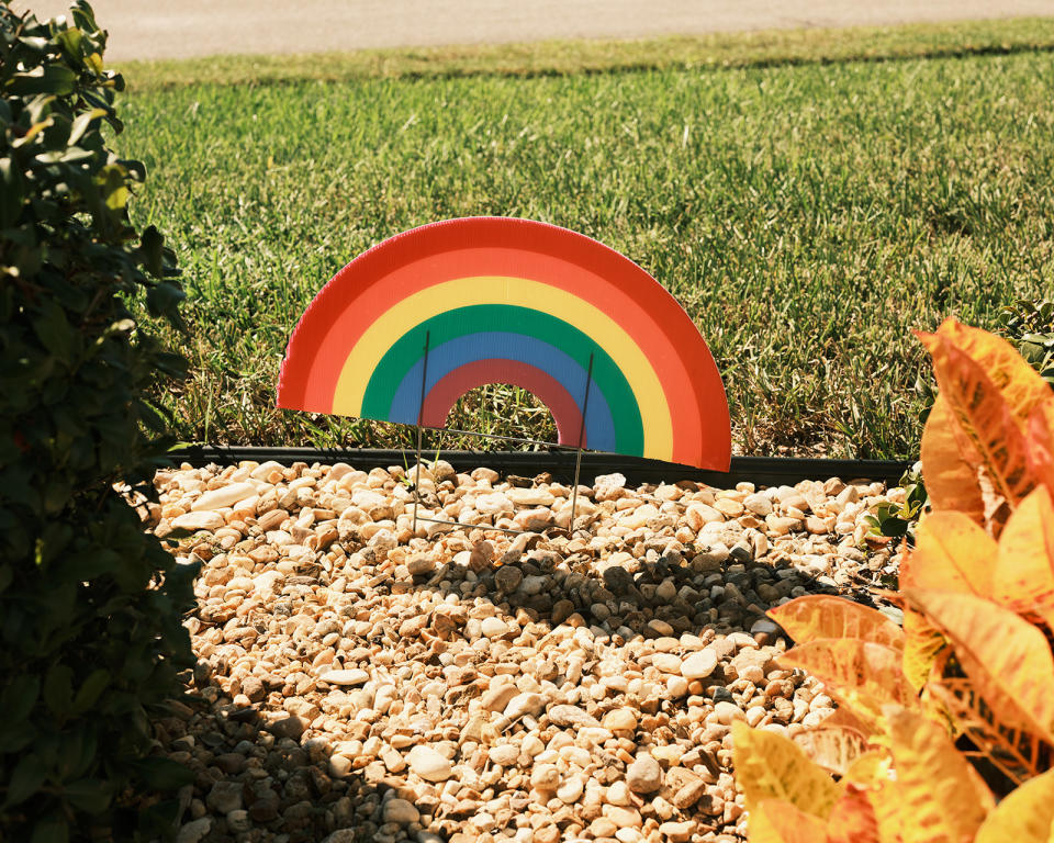 a carboard rainbow in the garden bed outside of Gail Foreman's house. There is green grass and the garden bed is comprised of stones.