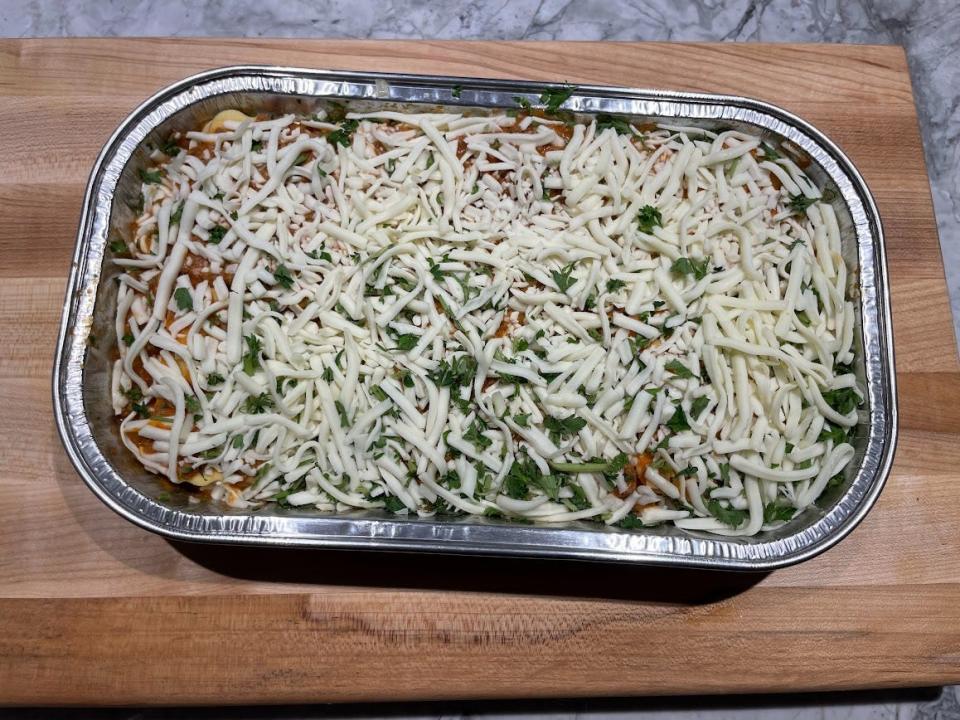 Tray of unheated ravioli lasagna with cheese shreds on top. The tray sits on a wooden cutting board