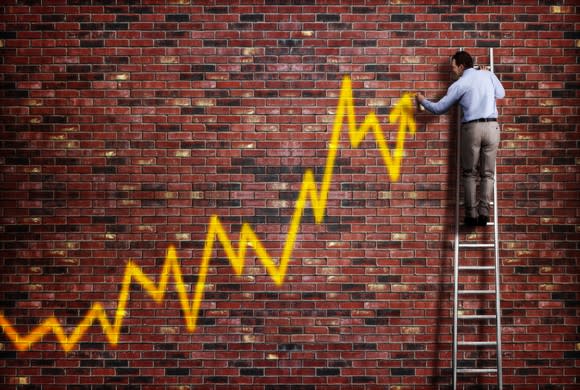 Man on ladder drawing a chart on a brick wall showing volatile stock gains.