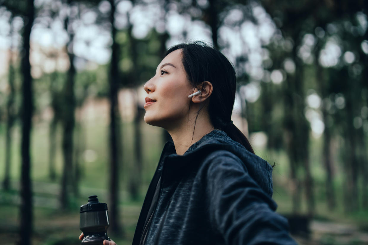 Confident and determined young Asian sports woman with wireless earphones holding a water bottle, getting ready to work out outdoors in nature park. Health and fitness training routine. Healthy living lifestyle