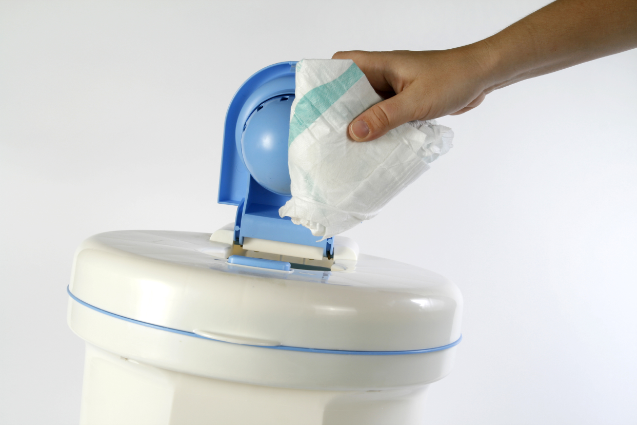 Hand putting used diaper into a white diaper pail with a blue top, at an angle, against a white background