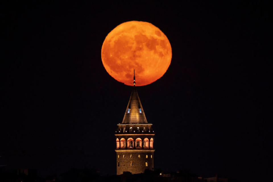 a blood orange color full moon rises behind a spire on the top of a large tower against a black sky.