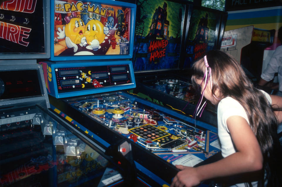 Person playing a vintage pinball machine at an arcade, with a Pac-Man-themed display visible above