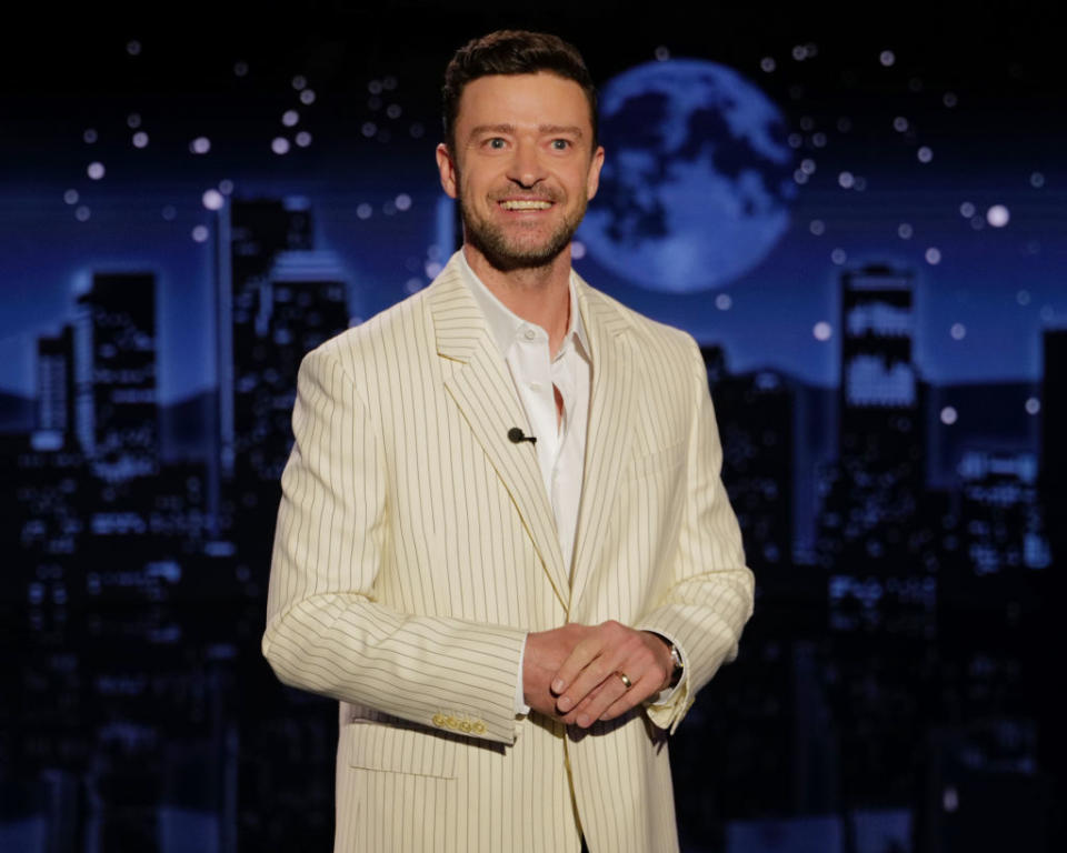 Justin Timberlake in a pinstripe suit and open-collar shirt, smiling onstage with a cityscape backdrop