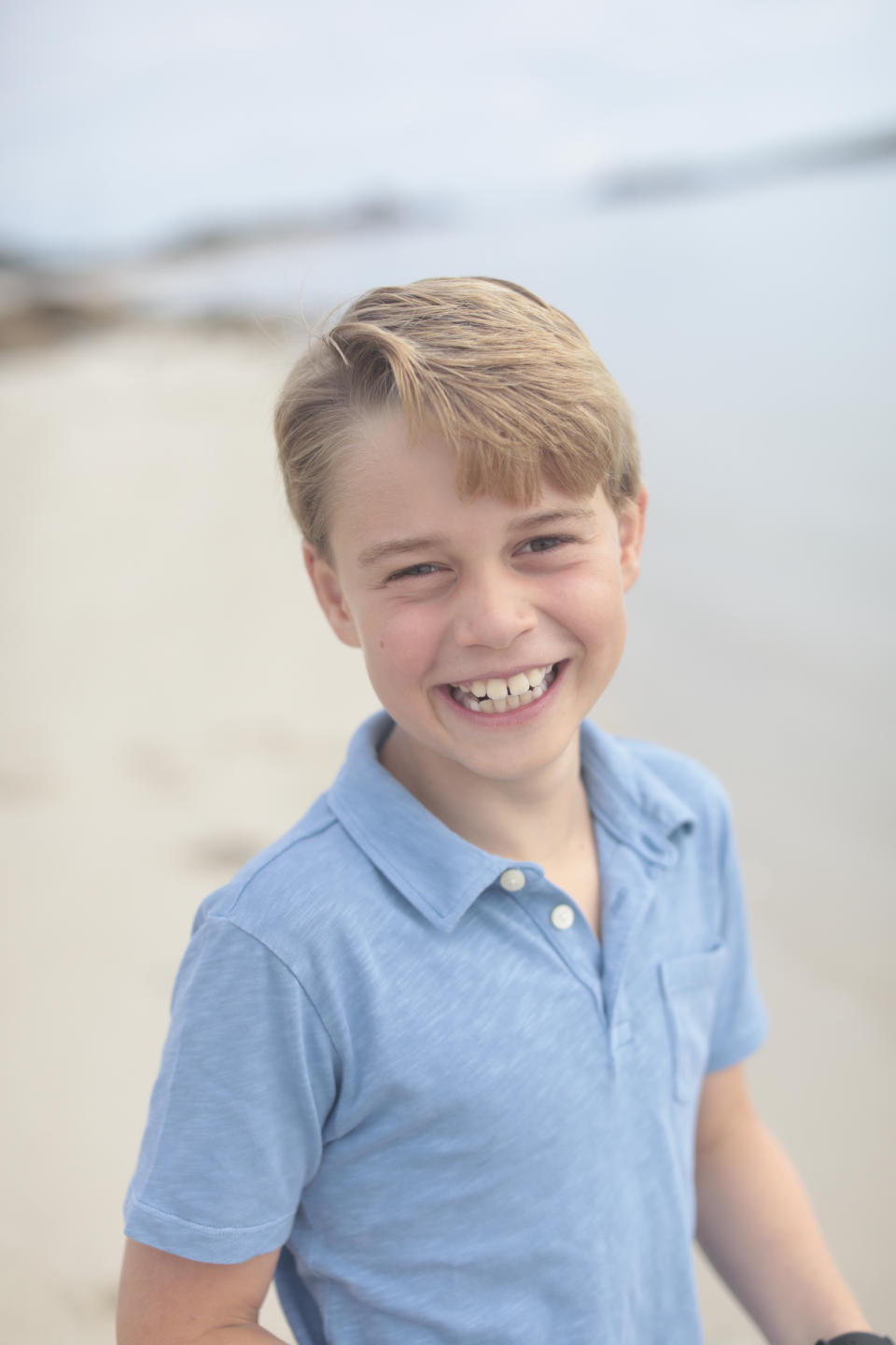 Prince George smiling on a beach wearing a blue polo shirt