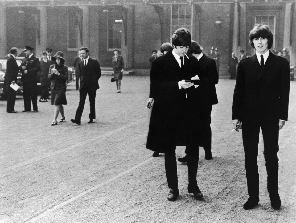 Paul McCartney and George Harrison at Buckingham Palace to receive their MBEs from the Queen, October 1965. Their manager Brian Epstein is in the background being interviewed by Maureen Cleave for the Evening Standard - Photo by H V Drees/Evening Standard/Getty Images