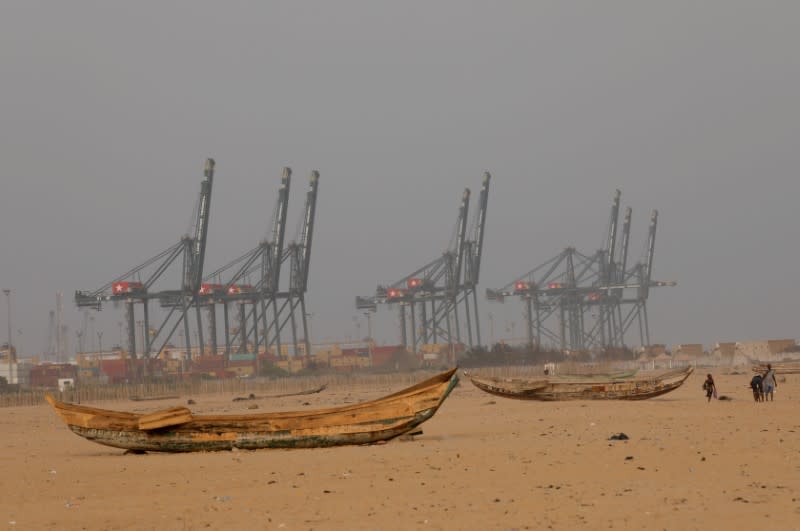 People walk past boats abandoned on the beach where the port of Lome can be seen in the distance in Lome
