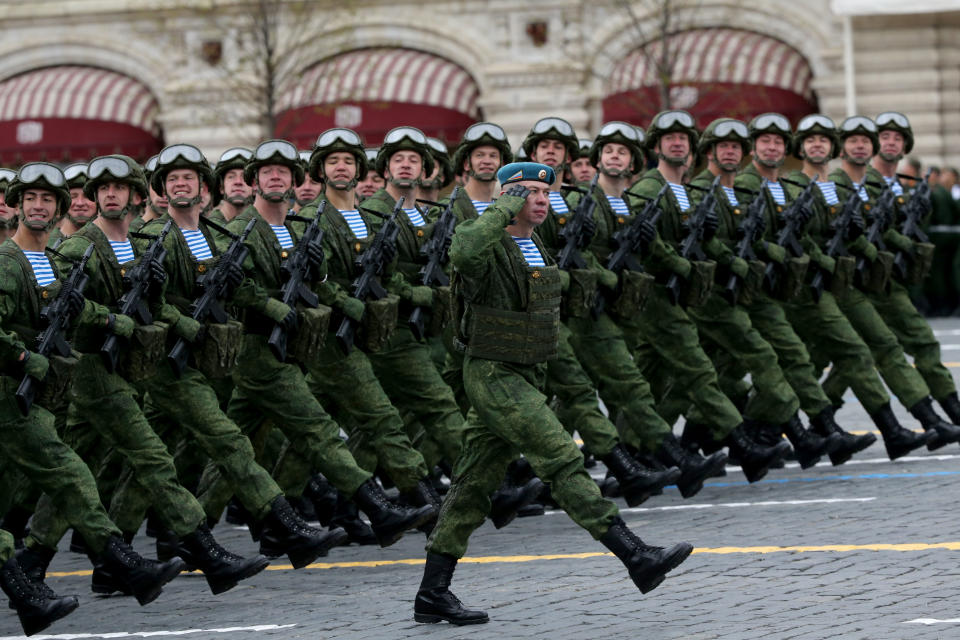 Several dozen soldiers wearing green uniforms and helmets with raised goggles and holding rifles march in lockstep on a road.