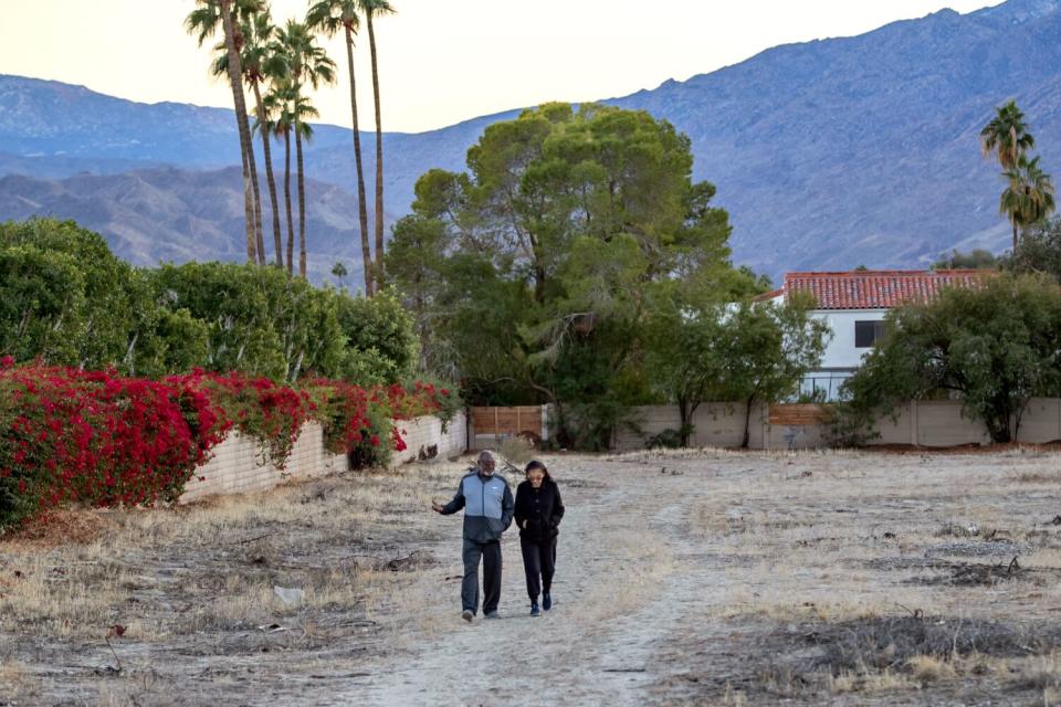 A couple walks across an arid lot of land flanked by greenery and palm trees with the mountains on the horizon.