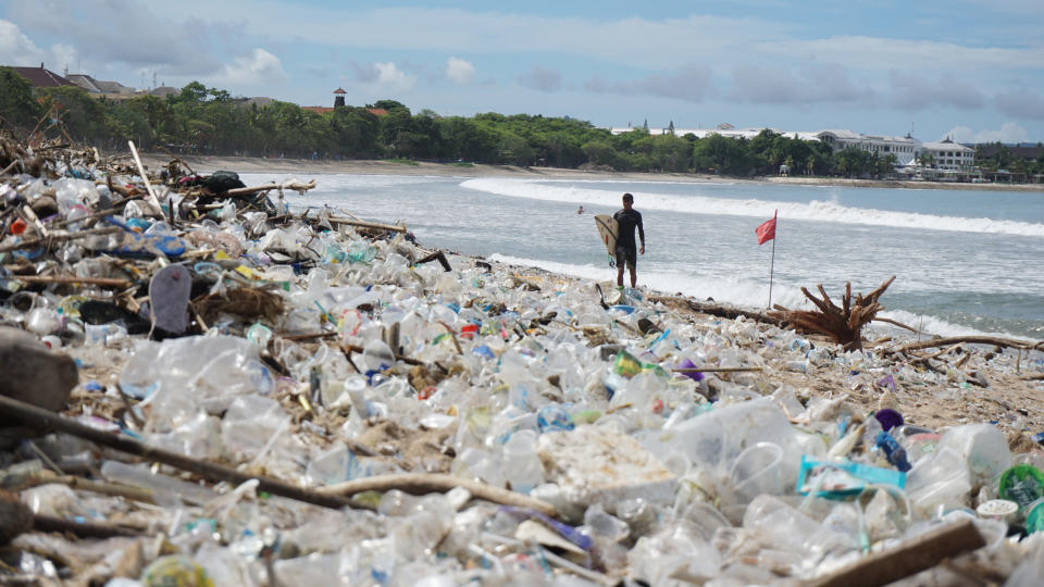 A surfer stands on the sand where a huge amount of plastic bottles and other rubbish sits on a Bali beach. 