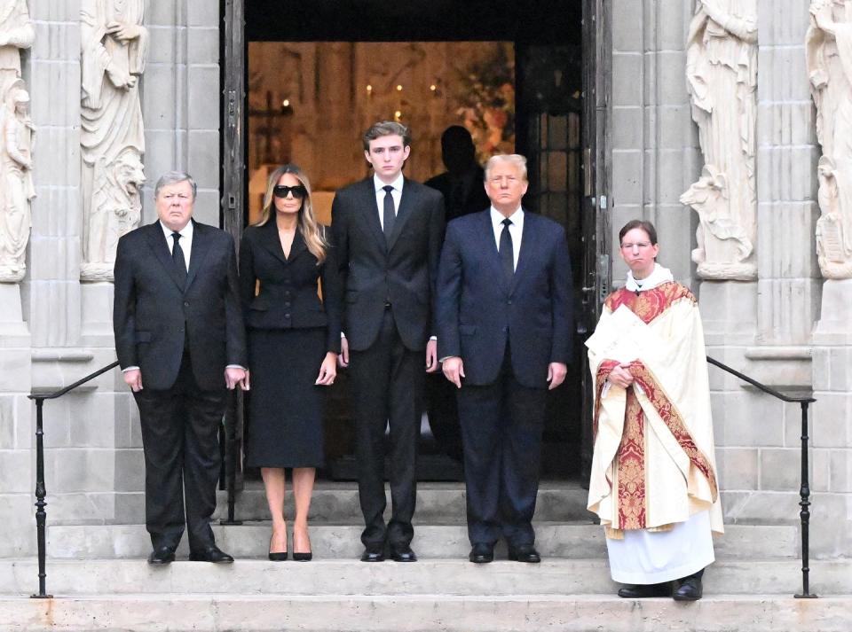 melania trump, barron trump, and donald trump stand on steps in front of an open set of doors, melania wears a dark skirt suit and sunglasses, barron and donald wear suits with ties and white collared shirts