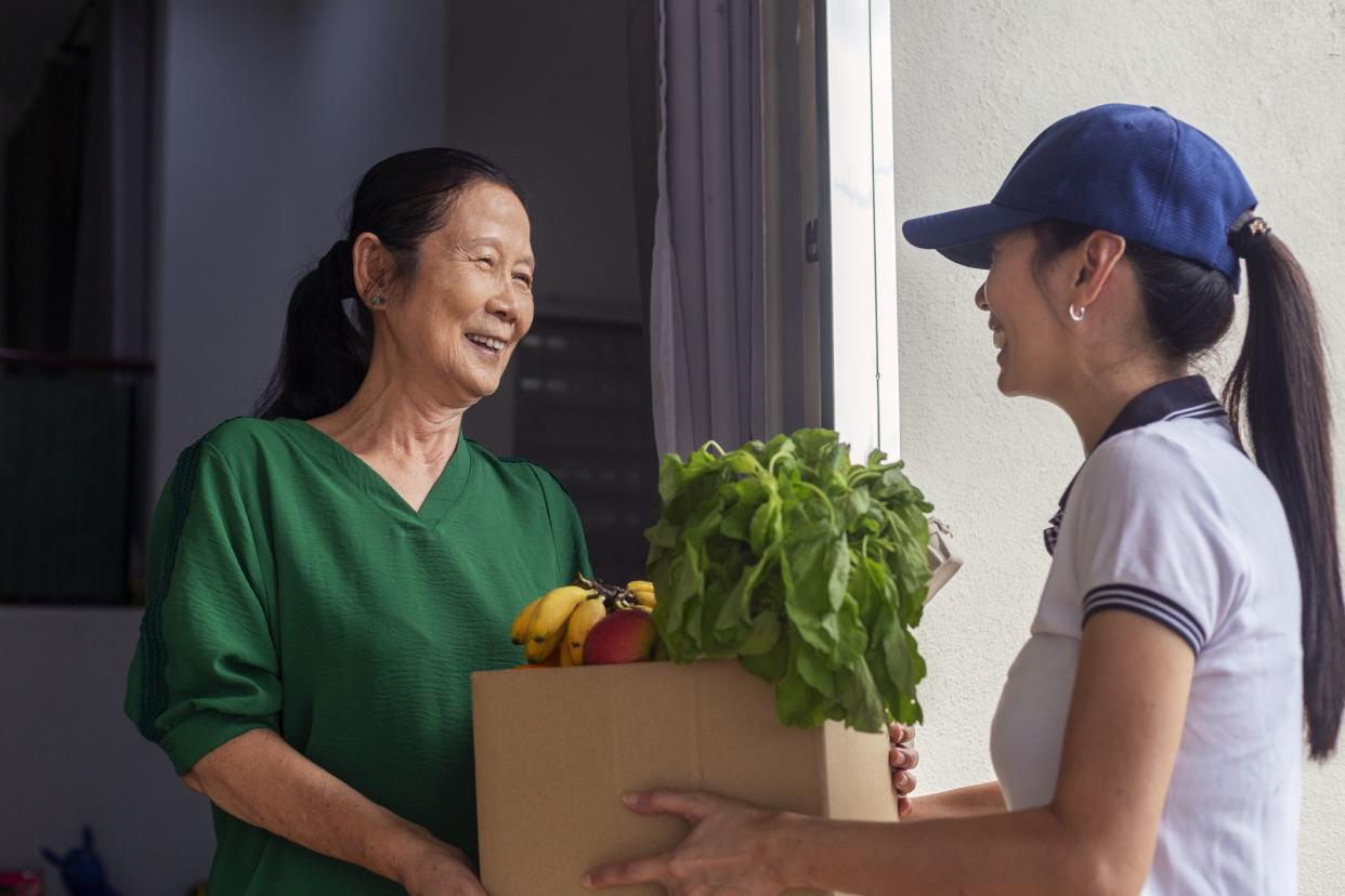 Asian deliver woman handling box of food, fruit, vegetable give to elderly female costumer in front of the house.