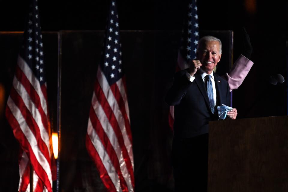 Joe Biden stands at a podium smiling in front of American flags, raising his right fist and holding his face mask with his left.