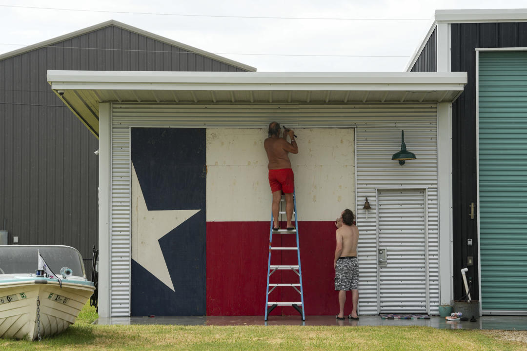 A man up on a ladder finishes up boarding up a home as another man standing nearby watches. 