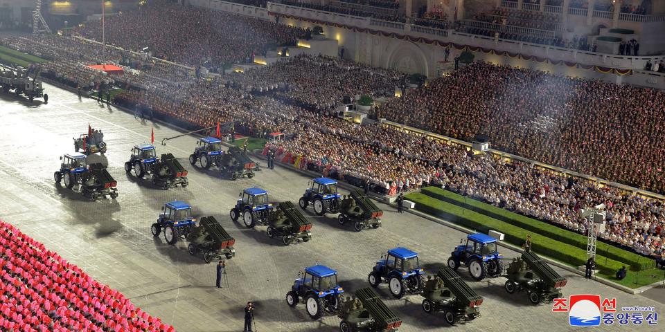 overhead shot of 9 blue tractors pulling rocket launchers before stadium packed with people