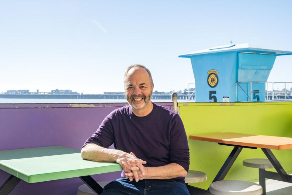 Ken Doctor sits at an outdoor table with the Santa Cruz wharf in the background.