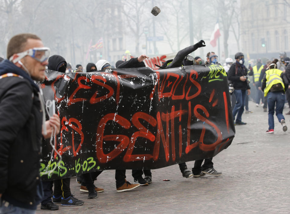 Youths throw a cobblestone on police officers, Saturday, March 16, 2019 in Paris. French yellow vest protesters clashed Saturday with riot police near the Arc de Triomphe as they kicked off their 18th straight weekend of demonstrations against President Emmanuel Macron. (AP Photo/Christophe Ena)