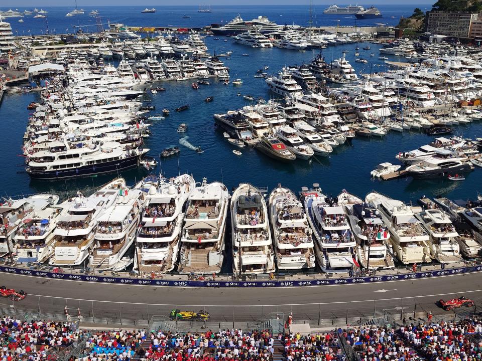 Grand Prix race track, with red and yellow cars on it, in front of the deep-blue French Riviera, filled with boats and yachts