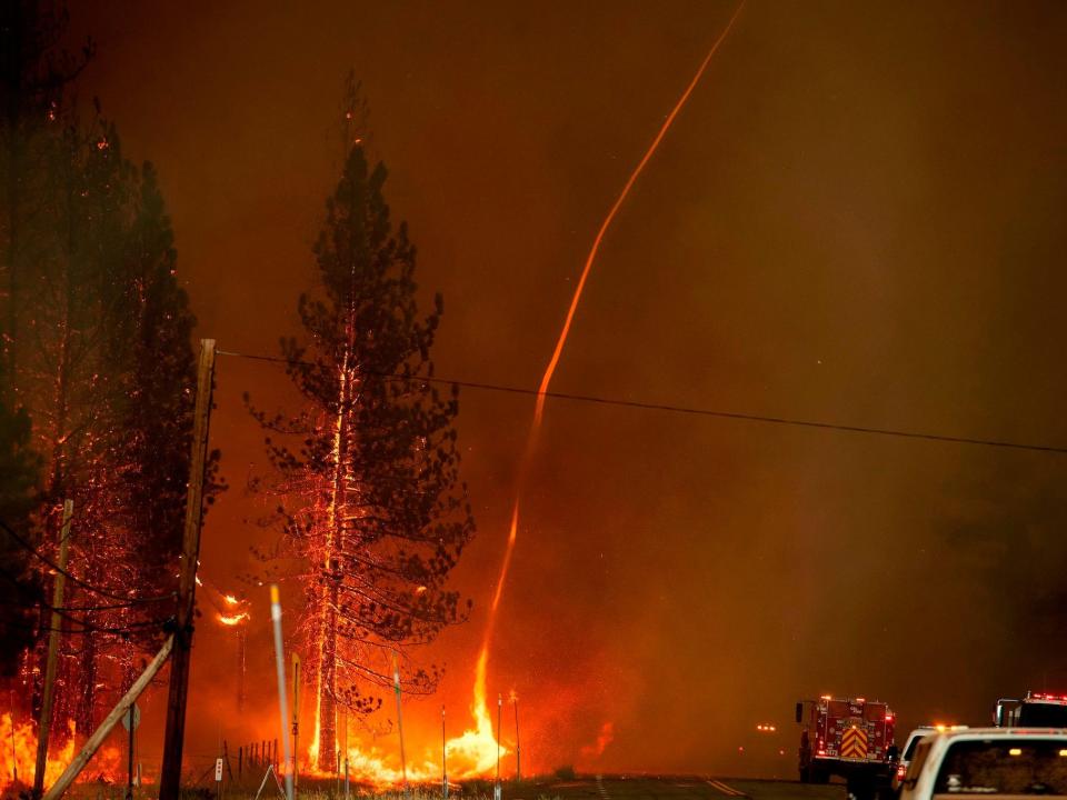 A fire whirl is seen in the midst of a wildfire, shooting a long strand of fire towards the sky.