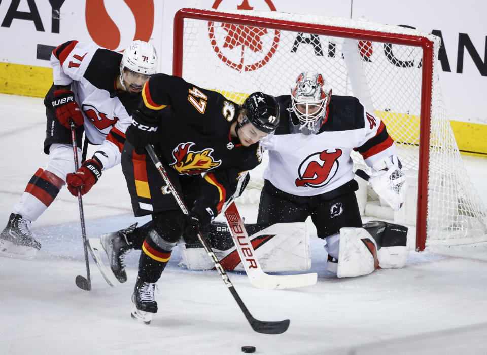 New Jersey Devils goalie Vitek Vanecek, right, guards the net as Calgary Flames forward Connor Zary (47) chases a loose puck while defenseman Jonas Siegenthaler, left, looks on during the second period of an NHL hockey match in Calgary, Alberta, Saturday, Dec. 9, 2023. (Jeff McIntosh/The Canadian Press via AP)