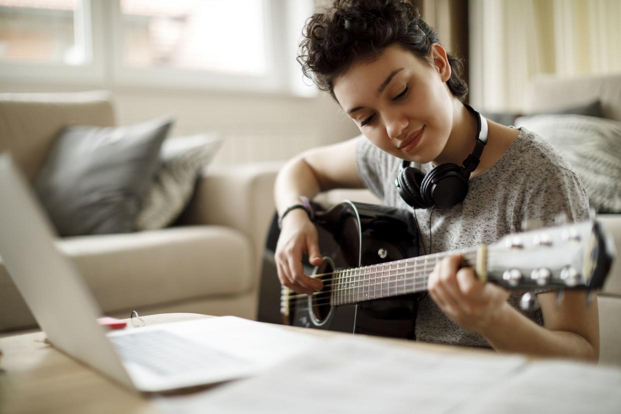young woman learning to play guitar, laptop open in front of her