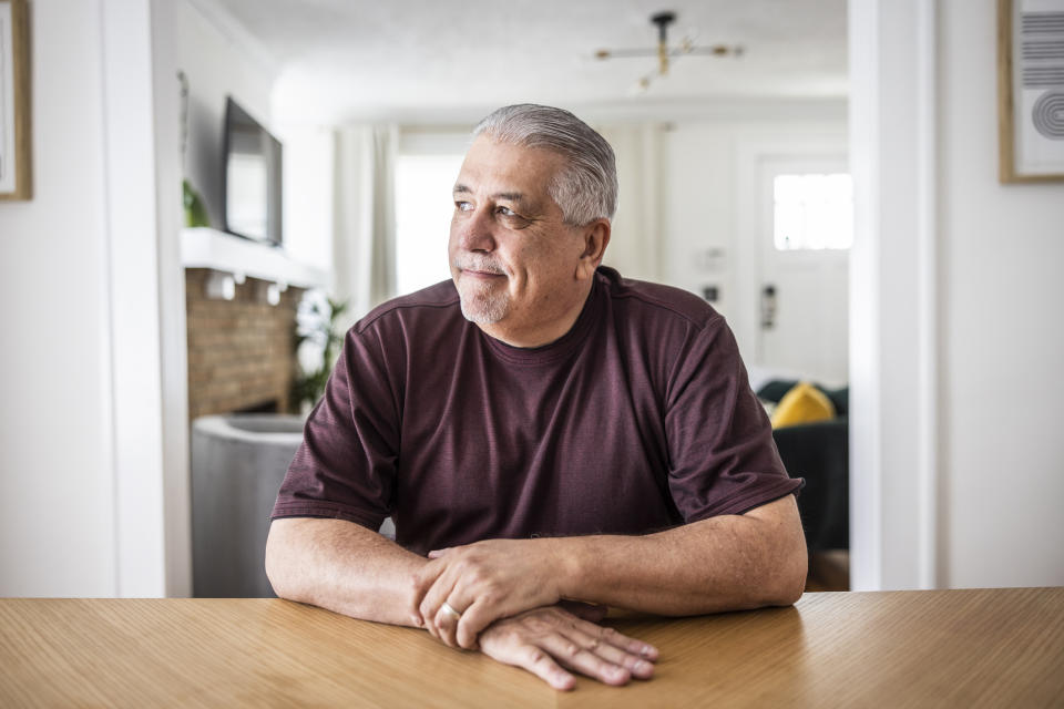 An older man sits at a wooden table, looking thoughtfully into the distance, inside a modern living room