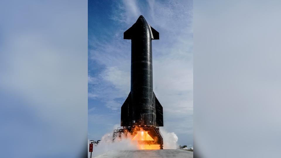     A sleek black rocket fires its engines while sitting on a launch pad. 