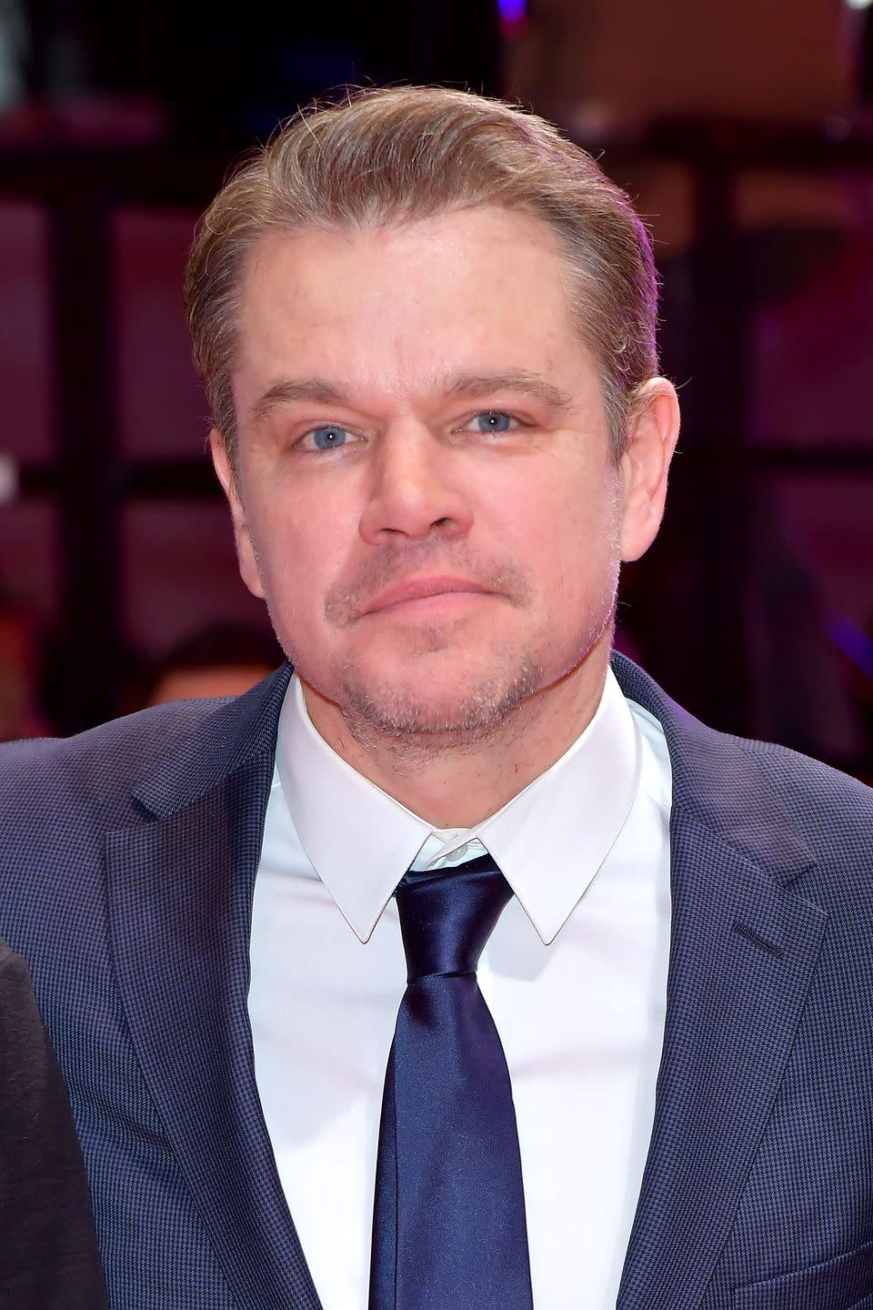 matt damon looks at the camera, he wears a blue suit jacket and tie with a white collared shirt