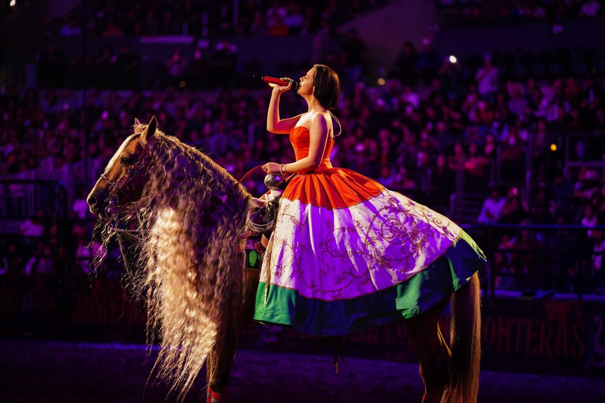 In her hometown of Los Angeles, Ángela Aguilar performs on horseback while donning a dazzling charro-style skirt resembling the Mexican flag and a red corset. Known for modernizing traditional charro outfits, the singer-songwriter expresses her love for her Mexican culture through her fashion.
