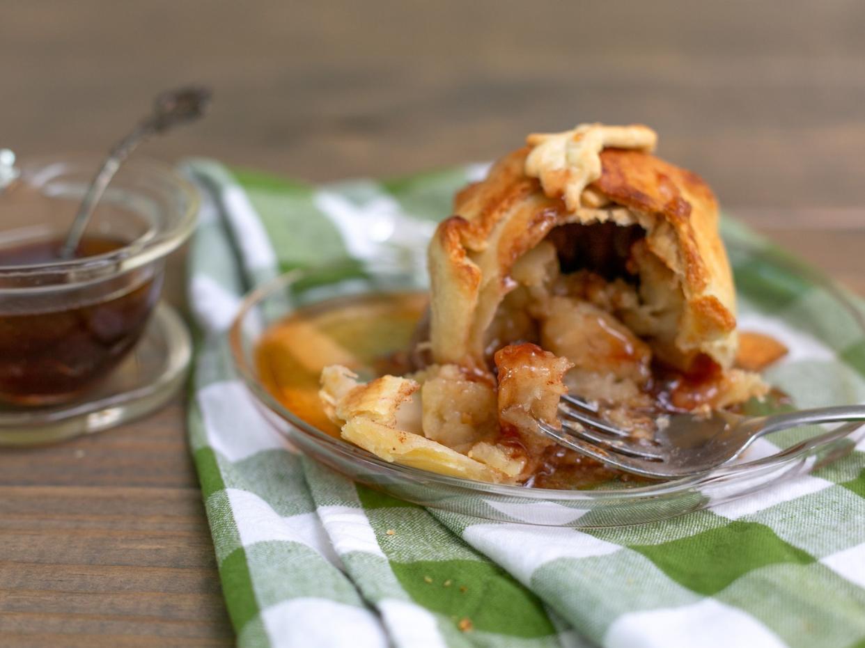 Homemade apple dumpling dessert presented on an apple-shaped plate with a green and white checked napkin on a wood table. A small apple-shaped bowl of cinnamon sugar syrup in the background.