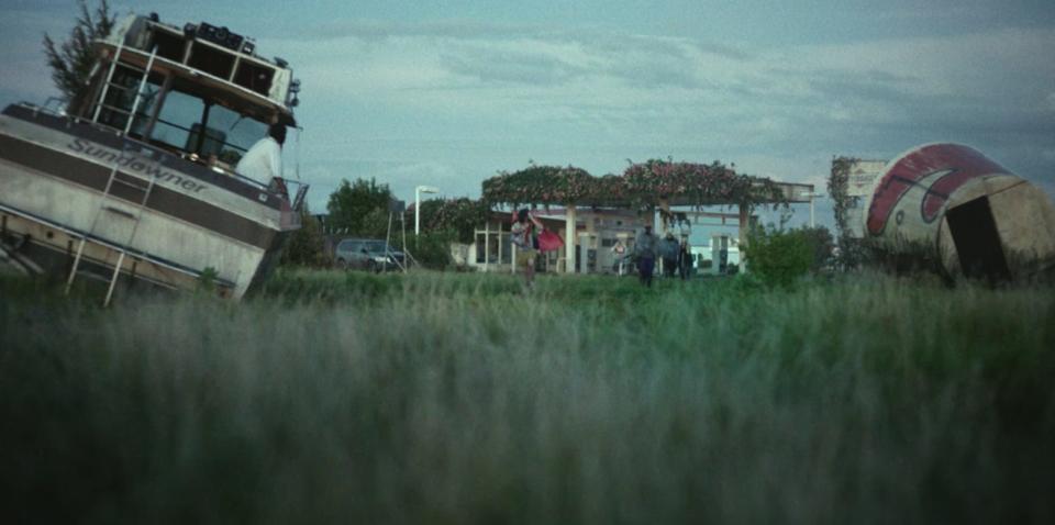 an overgrown gas station, non-functional and covered on the top with vibes. to the left, in the foreground, there's a grounded boat with plants sprouting out of it. to the right, there's a giant, overturned bucket of fried chicken — a sculpture, as if for a fast food joint