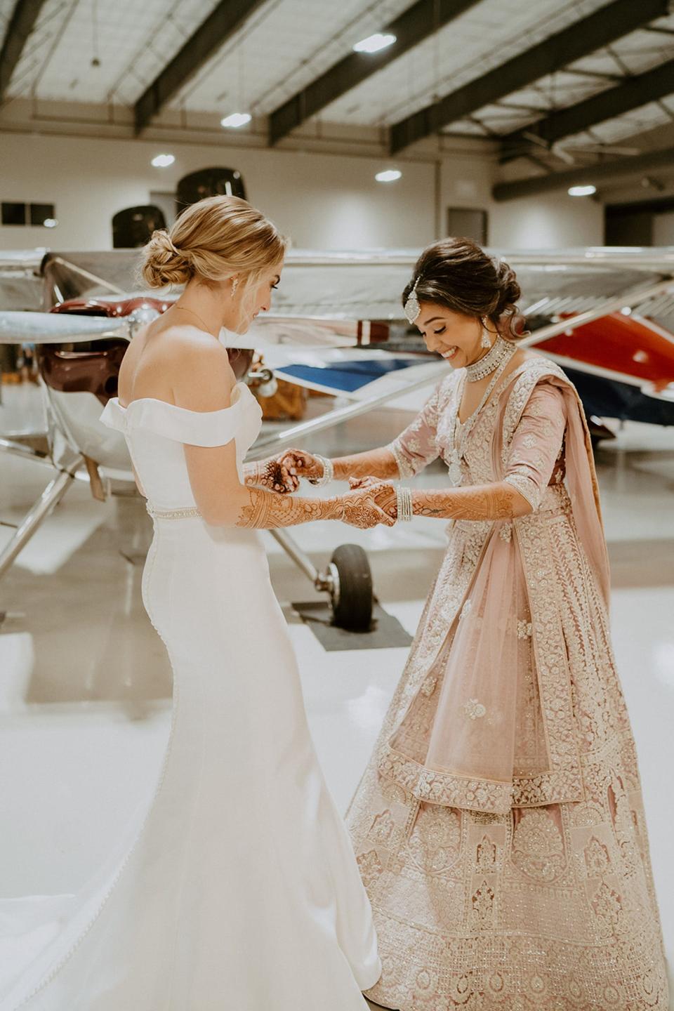 Two brides hold hands and look at each other's wedding outfits in front of planes.