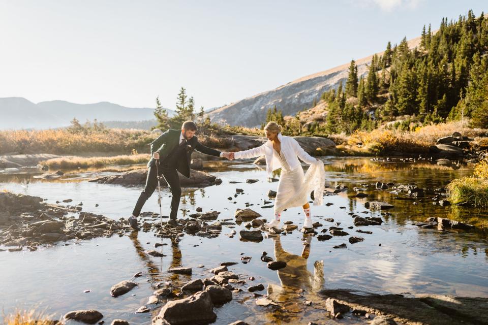 A couple holds hands in their wedding attire as they walk across a rocky pond.