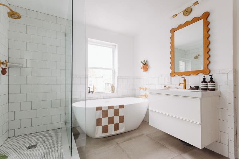 Philadelphia residence with white walls, lots of wood details: bathroom with oval freestanding tub, brown and white checkered bath mat, glass shower with white tiles, rose gold hardware, plants. Window over tub and in shower, white sink and cabinet with wavy frame mirror