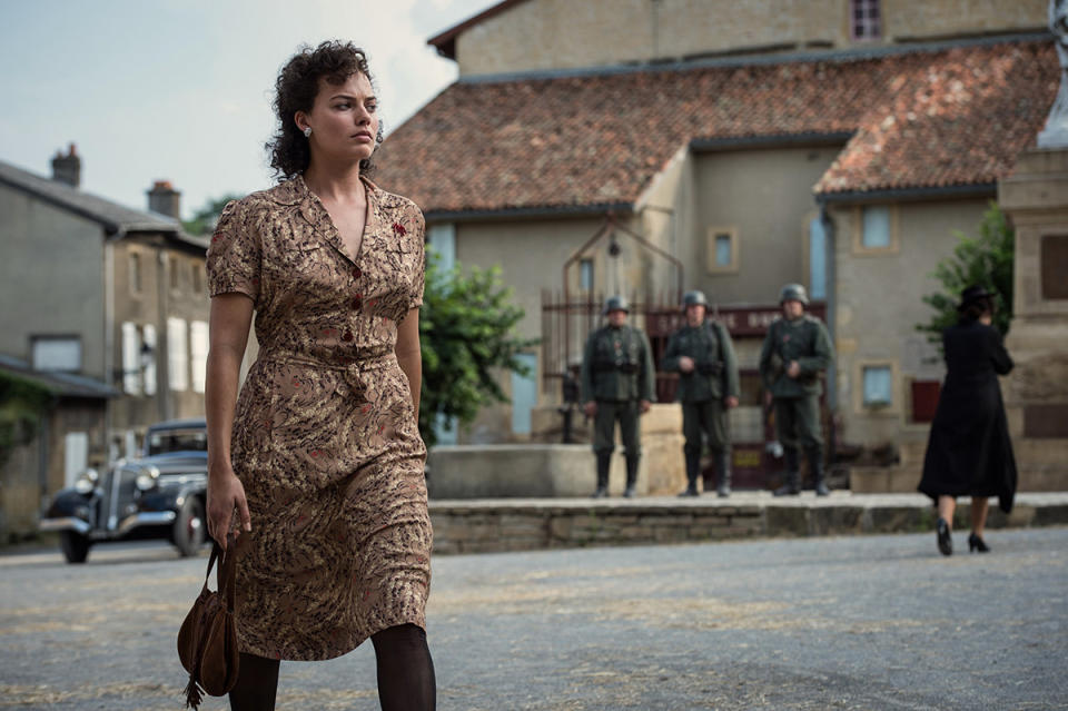 Margot Robbie walks across a town square in a dark wig