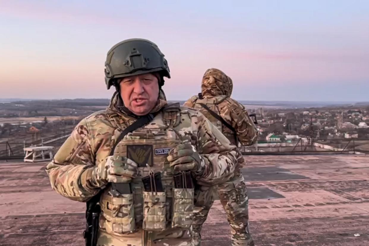 Yevgeny Prigozhin, in camouflage and helmet, on the top of a tall building looking over a town, with another combatant in uniform behind him.