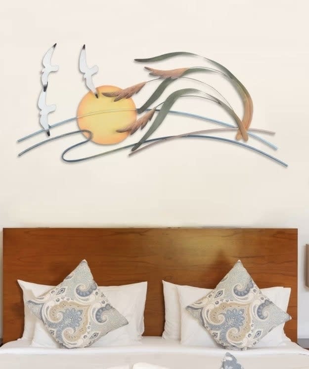 A bed with two pillows featuring decorative patterns, with a wall-mounted metal art piece above portraying birds, reeds, and a sun scene