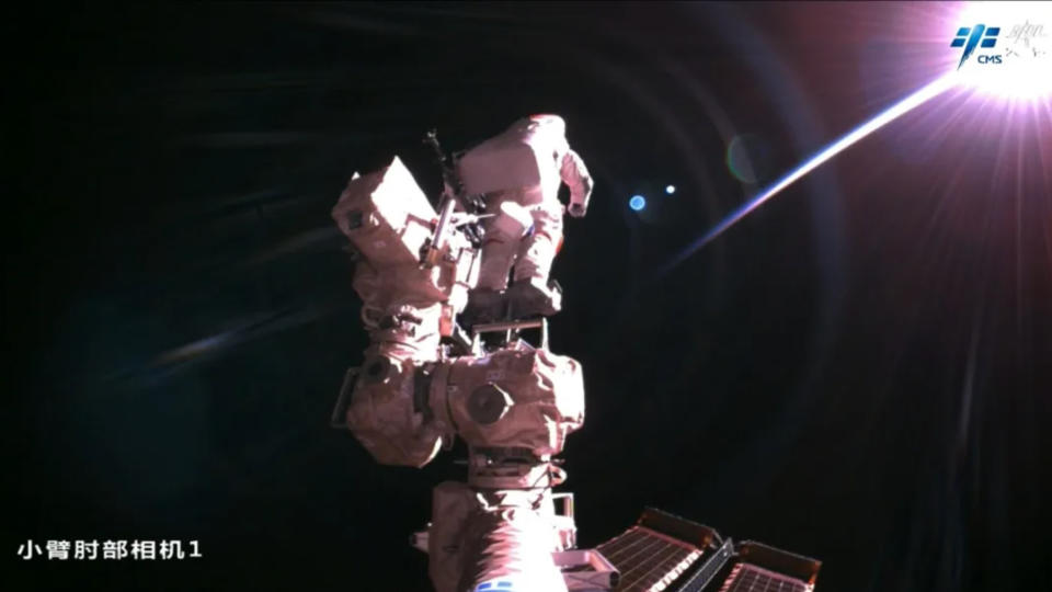 an astronaut in a white spacesuit stands at the end of a robotic arm with the blackness of space in the background