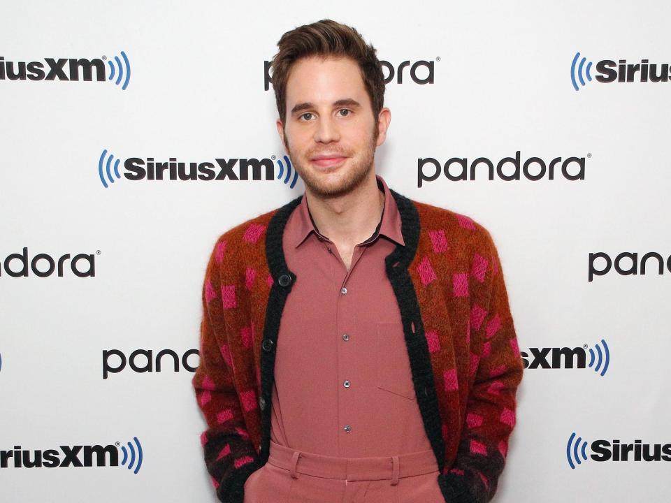 Ben Platt at an event. He wears a red checkered jacket and a salmon pink jumpsuit.