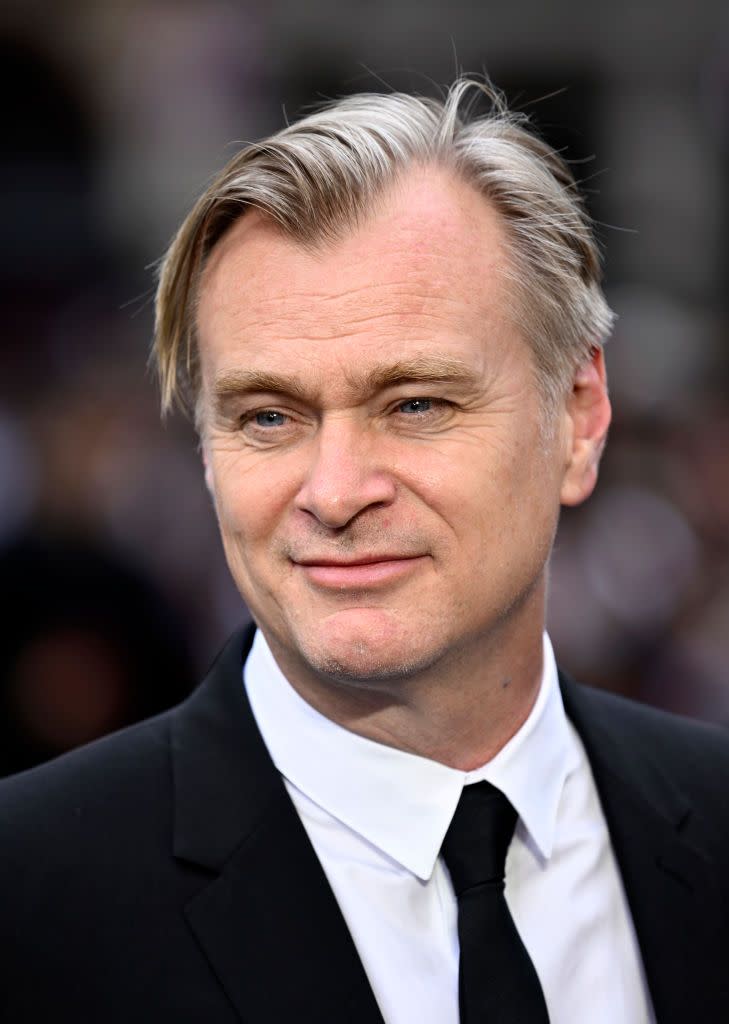 christopher nolan smiles and looks to the left, he wears a black suit jacket and tie with a white collared shirt