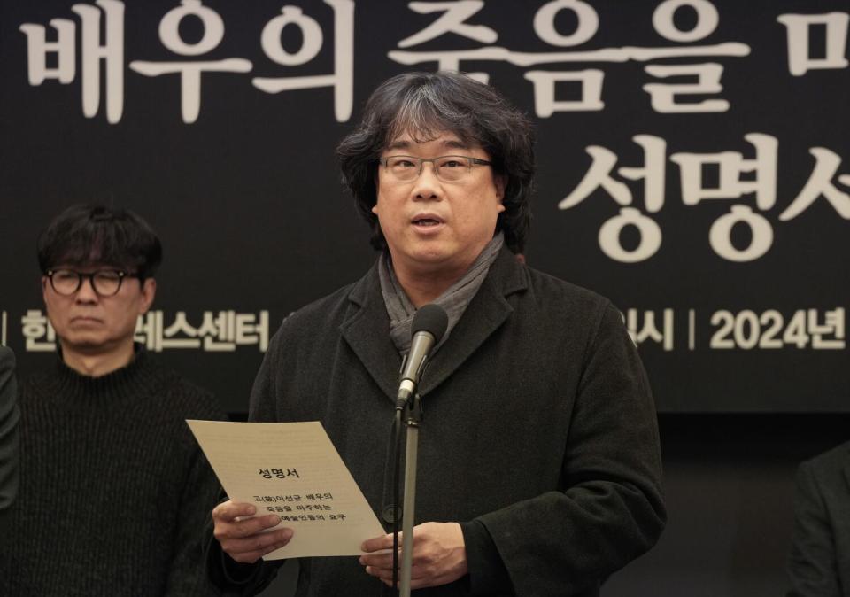 A man with dark hair, wearing glasses and dark clothes, holds a sheet of paper while standing before a microphone