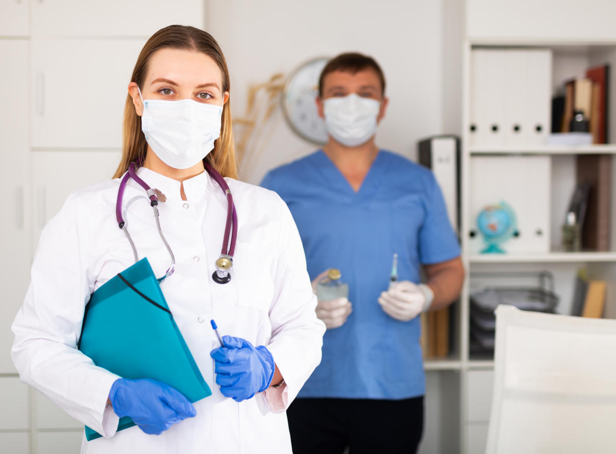 Young woman general practitioner in white lab coat and face mask standing in clinic, holding clipboard with medical records